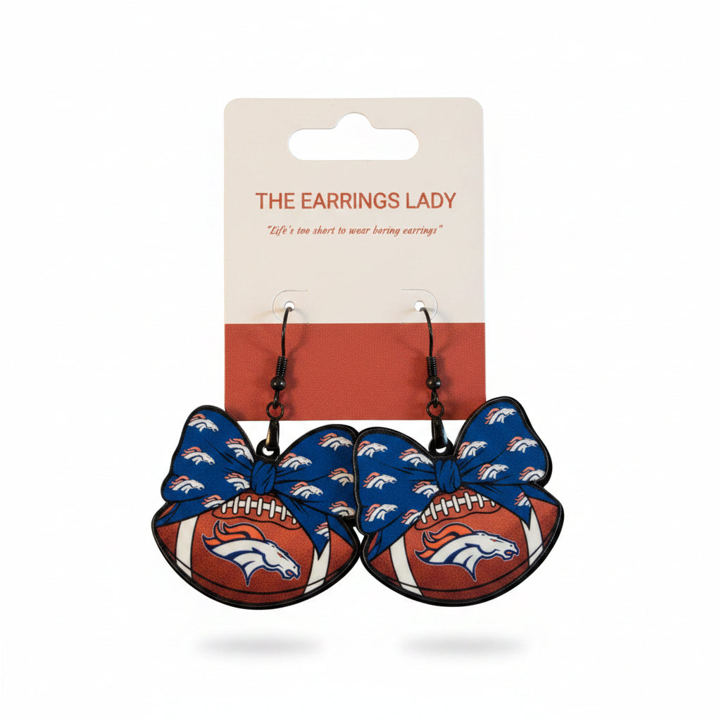 A pair of Denver Broncos themed dangling earrings held in a hand. The earrings are shaped like football-shaped bows, featuring the team's blue and orange colors with the horse logo.