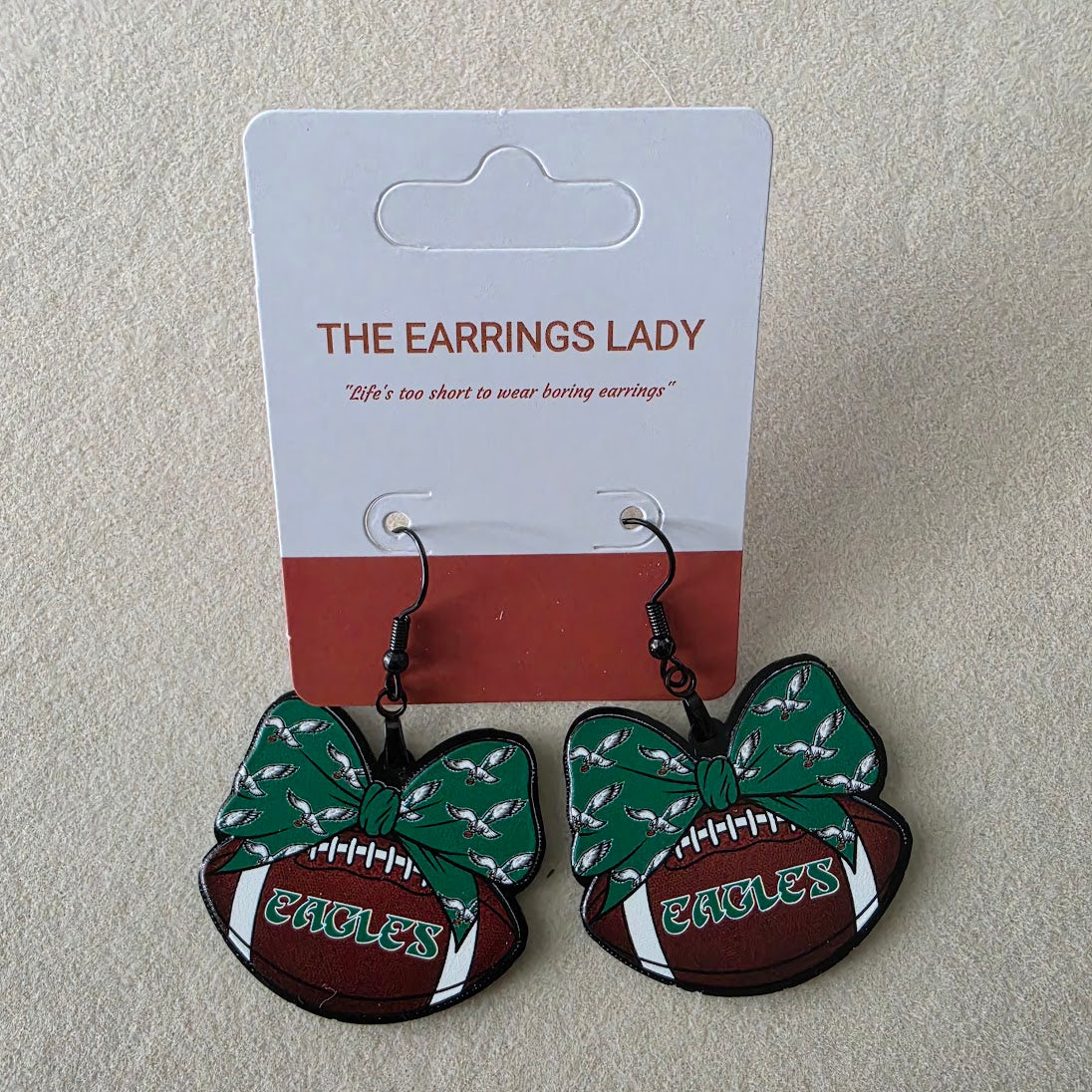 A pair of Eagles-themed dangling earrings displayed on their original white and red backing card, which is branded 'THE EARRINGS LADY'. The earrings feature a green patterned bow, a brown football, and white accents with 'EAGLES' text.
