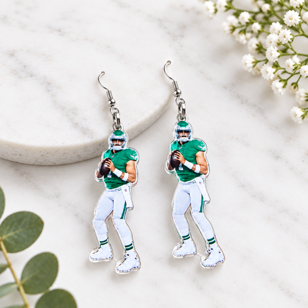 A pair of football player dangle earrings on a white and orange card from 'The Earrings Lady', placed on a wooden table. The earrings feature a detailed graphic of a football player in a green jersey and white pants.