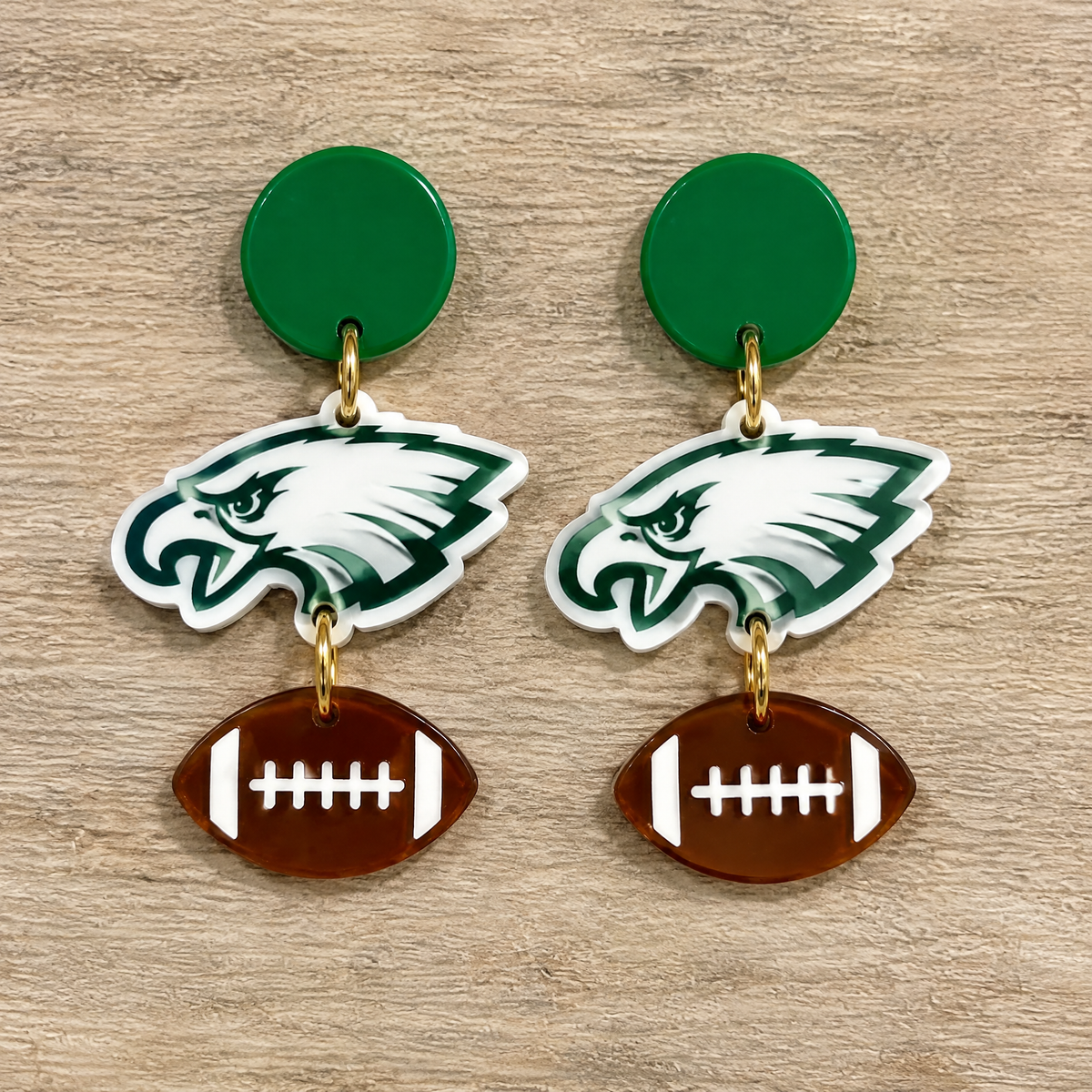 A pair of Philadelphia Eagles themed drop earrings on a light wood background. Each earring features a green circular stud, a white and green Eagles head logo charm, and a brown football charm with white laces.