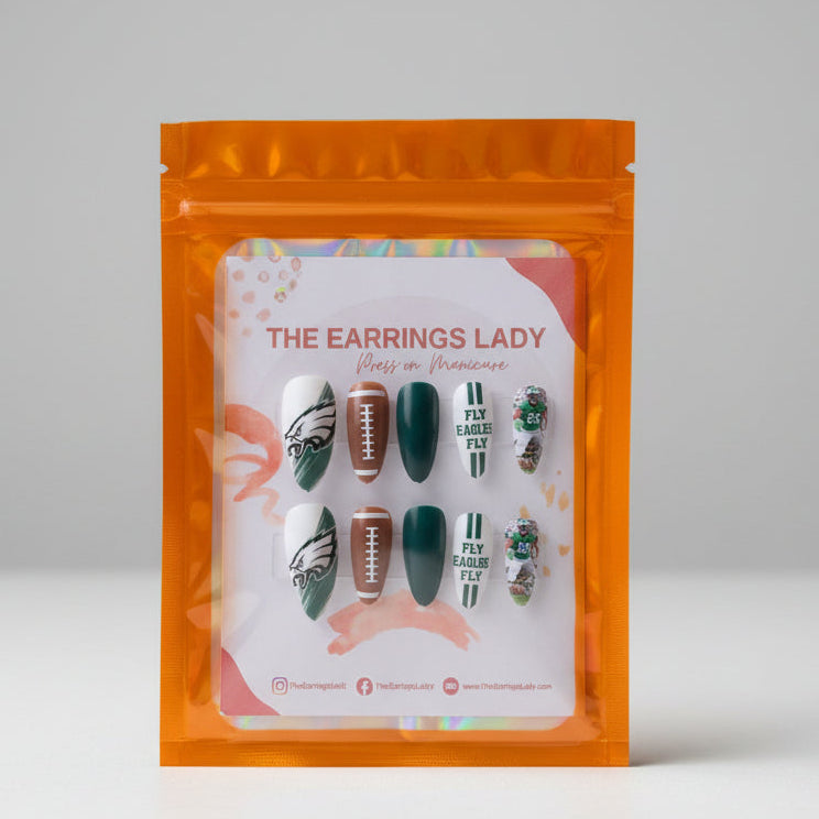 A package of press-on nails displayed inside an orange resealable pouch. The nails are decorated with Philadelphia Eagles-themed designs, including green, white, brown, and striped patterns.
