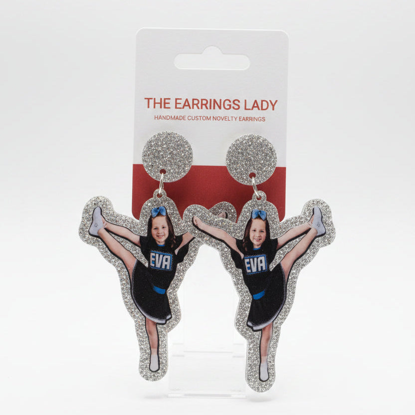 A hand holds a pair of custom cheerleader-shaped stud earrings on their display card, which reads 'THE EARRINGS LADY'. The earrings feature a printed photo of a cheerleader in a black and blue uniform surrounded by silver glitter.