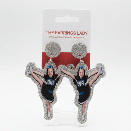 A hand holds a pair of custom cheerleader-shaped stud earrings on their display card, which reads 'THE EARRINGS LADY'. The earrings feature a printed photo of a cheerleader in a black and blue uniform surrounded by silver glitter.