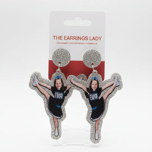 A hand holds a pair of custom cheerleader-shaped stud earrings on their display card, which reads 'THE EARRINGS LADY'. The earrings feature a printed photo of a cheerleader in a black and blue uniform surrounded by silver glitter.