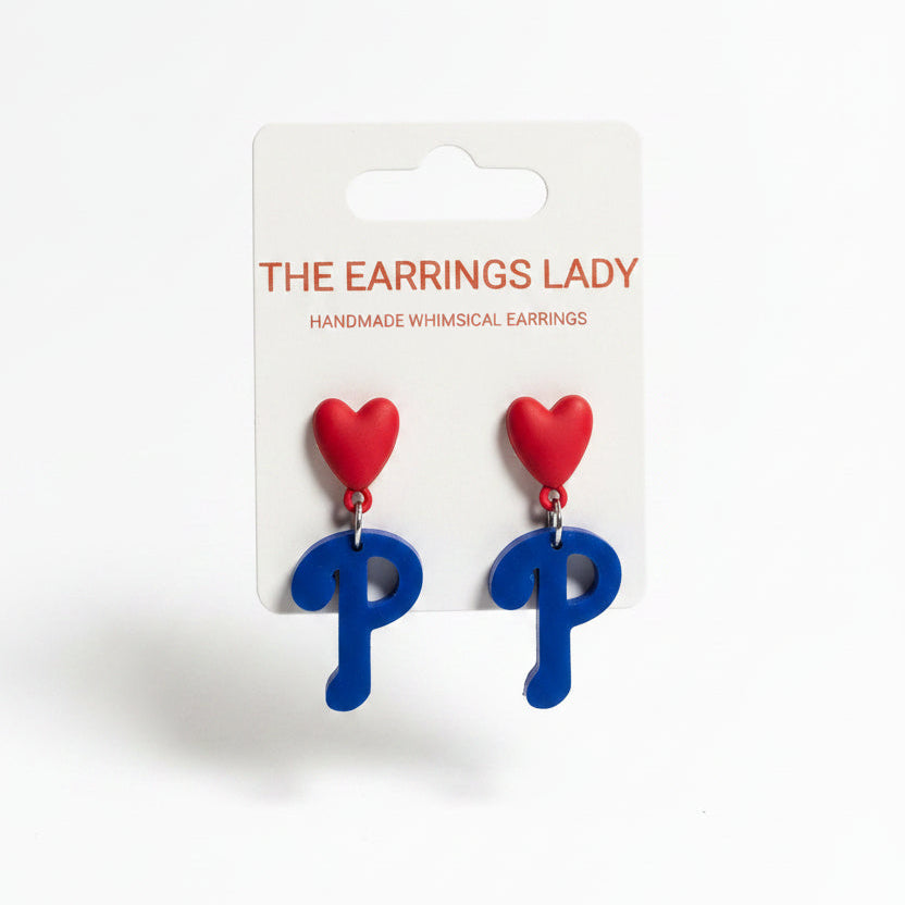 A hand holds a card displaying a pair of whimsical earrings shaped like the letter P, with a red heart on top and a blue body. The background shows a jewelry organizer filled with small parts and other earring cards.