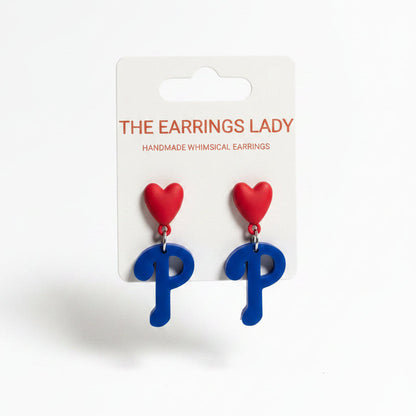 A hand holds a card displaying a pair of whimsical earrings shaped like the letter P, with a red heart on top and a blue body. The background shows a jewelry organizer filled with small parts and other earring cards.