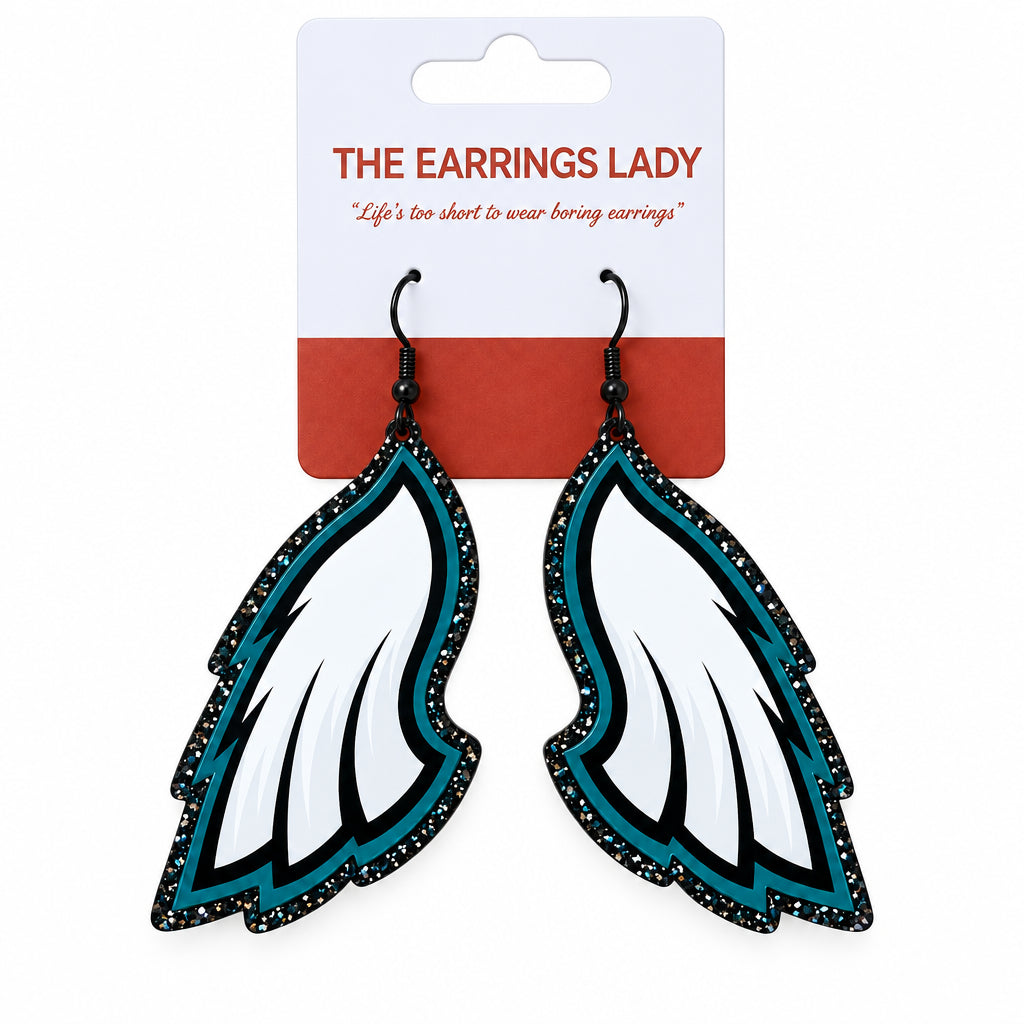 A pair of wing-shaped dangle earrings on a white and coral card from 'The Earrings Lady'. The earrings feature white feathers with black glittery edges and teal accents.