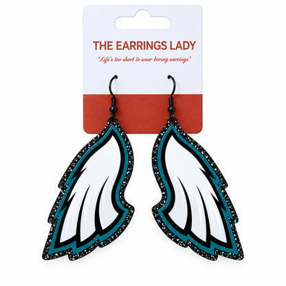 A pair of wing-shaped dangle earrings on a white and coral card from 'The Earrings Lady'. The earrings feature white feathers with black glittery edges and teal accents.
