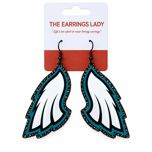 A pair of wing-shaped dangle earrings on a white and coral card from 'The Earrings Lady'. The earrings feature white feathers with black glittery edges and teal accents.