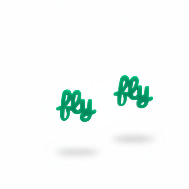 A pair of small green dangling earrings on a white and red blister card, photographed on a beige carpet with a dark textured surface at the right edge. The card reads 'THE EARRINGS LADY'.