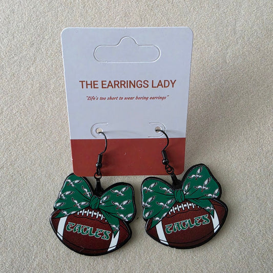 A pair of Eagles-themed dangling earrings displayed on their original white and red backing card, which is branded 'THE EARRINGS LADY'. The earrings feature a green patterned bow, a brown football, and white accents with 'EAGLES' text.