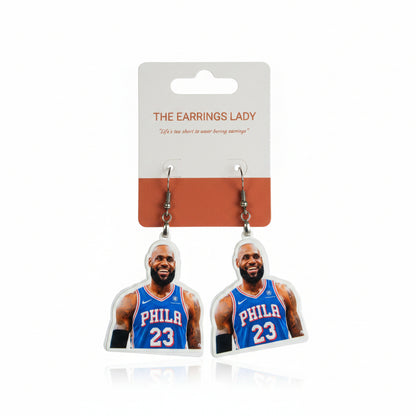 A pair of dangling earrings shaped like basketball player LeBron James in a blue Philadelphia 76ers jersey with the number 23, hanging from a white and orange card labeled 'THE EARRINGS LADY' above a black keyboard on a gray gridded cutting mat.