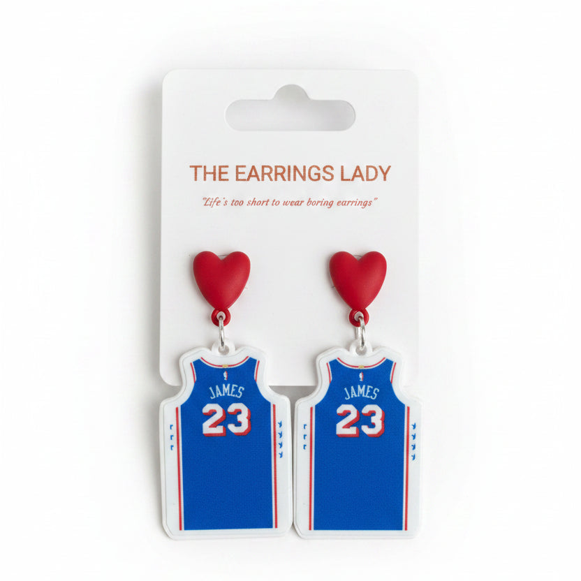 A pair of blue basketball jersey dangle earrings with red heart tops, displayed on a white card labeled 'THE EARRINGS LADY' and held by a hand.