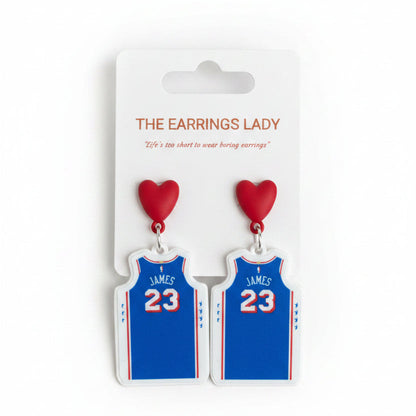 A pair of blue basketball jersey dangle earrings with red heart tops, displayed on a white card labeled 'THE EARRINGS LADY' and held by a hand.