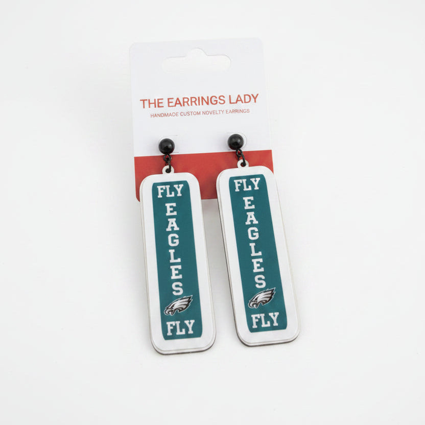 A pair of Philadelphia Eagles themed rectangular dangle earrings by The Earrings Lady, featuring a white and dark green design with the team logo and 'FLY EAGLES FLY' text, shown on their retail card against a gray tabletop.