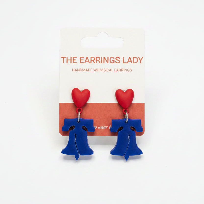 Handmade blue and red heart dangle earrings on a white backing card from 'The Earrings Lady', held by a hand in a jewelry-making workspace with small parts and pliers visible on the table.