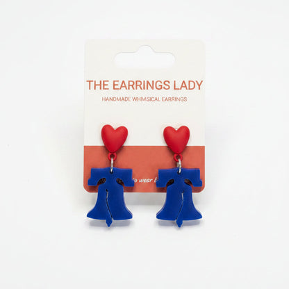 Handmade blue and red heart dangle earrings on a white backing card from 'The Earrings Lady', held by a hand in a jewelry-making workspace with small parts and pliers visible on the table.