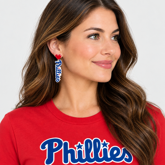 A close-up of a woman wearing a dangling earring with a red heart and the word 'Phillies' in white script outlined in blue, paired with a red Phillies t-shirt.