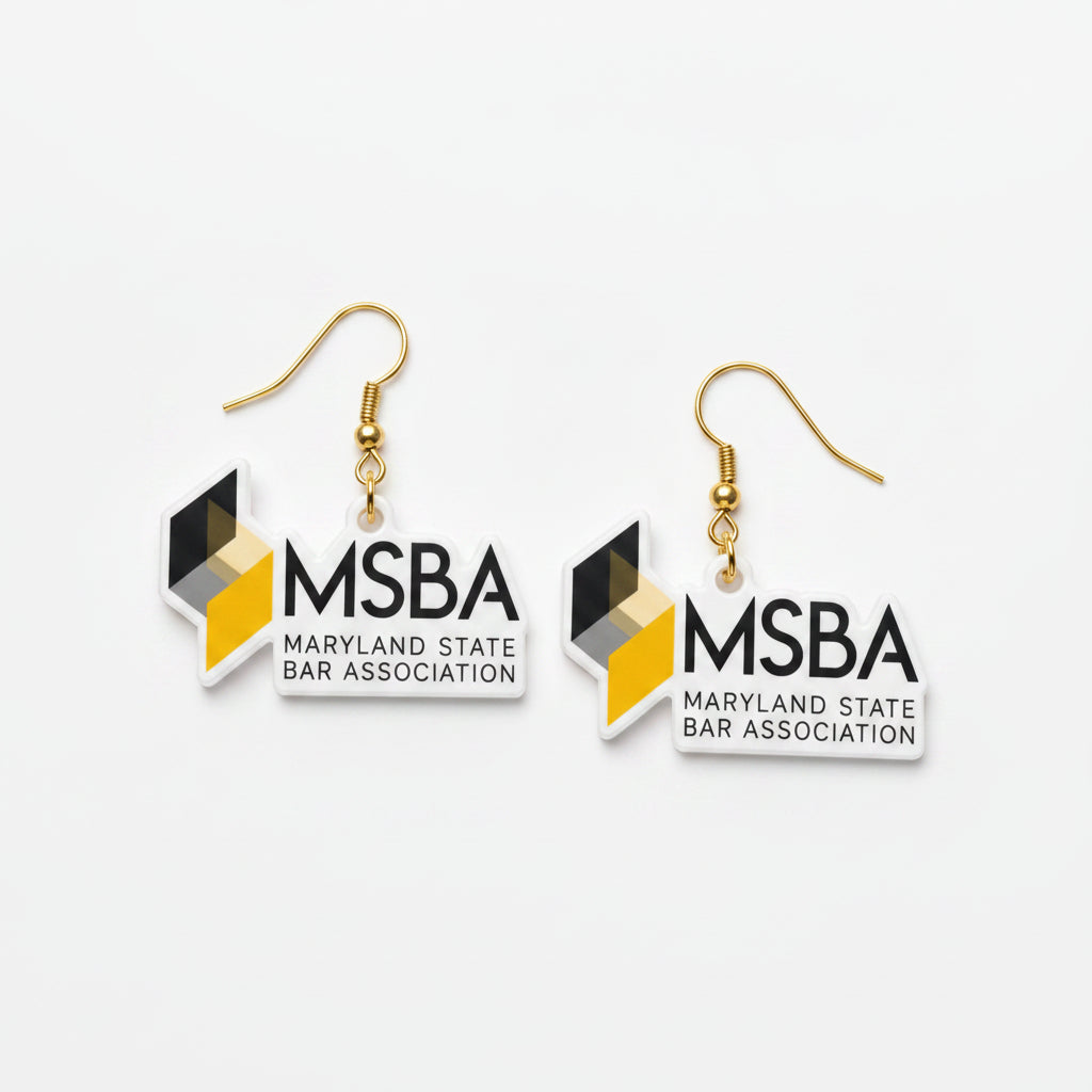 A pair of dangle earrings featuring the Maryland State Bar Association (MSBA) logo. The earrings have a white rectangular pendant with black text and a geometric logo in black, gray, and yellow, hanging from silver fishhook wires on a light gray background.