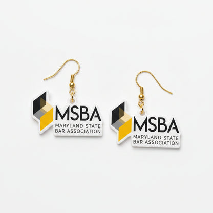 A pair of dangle earrings featuring the Maryland State Bar Association (MSBA) logo. The earrings have a white rectangular pendant with black text and a geometric logo in black, gray, and yellow, hanging from silver fishhook wires on a light gray background.