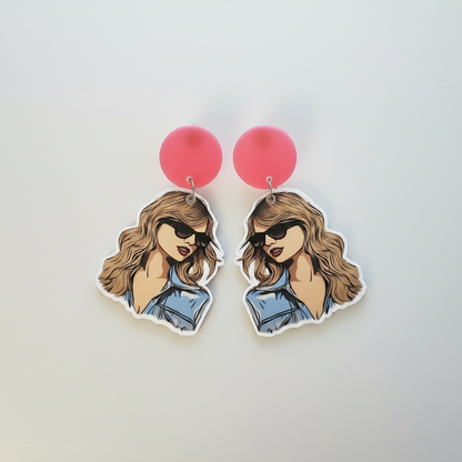 Earrings with cartoon-style illustration of a woman wearing sunglasses and a denim jacket on a light gray Taylor Swift inspired pop star portrait earrings.background.