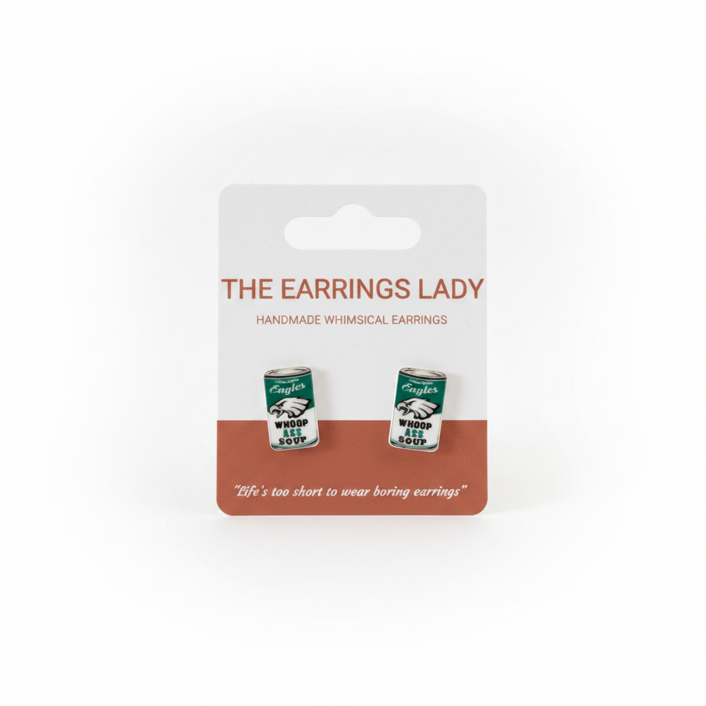 A pair of small tin-can-shaped earrings displayed on a white retail card labeled 'THE EARRINGS LADY'. A hand is holding the card in front of an orange pegboard background.