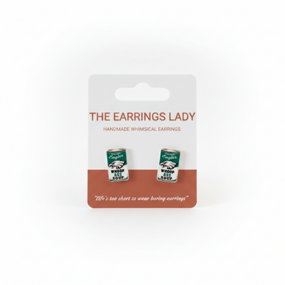 A pair of small tin-can-shaped earrings displayed on a white retail card labeled 'THE EARRINGS LADY'. A hand is holding the card in front of an orange pegboard background.